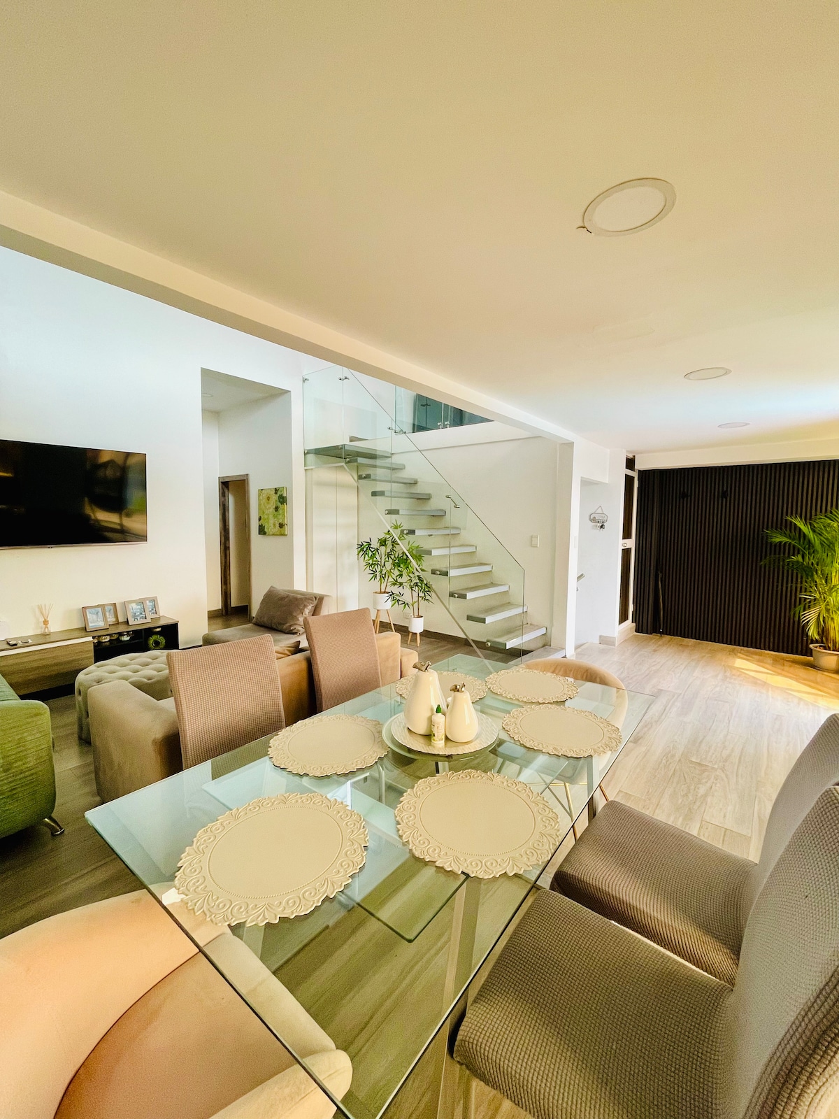 A dining area features a glass table set with decorative placemats. Soft seating is arranged around the table, leading to a staircase with a modern design. Natural light streams through the space, highlighting the greenery of a potted plant in the corner.