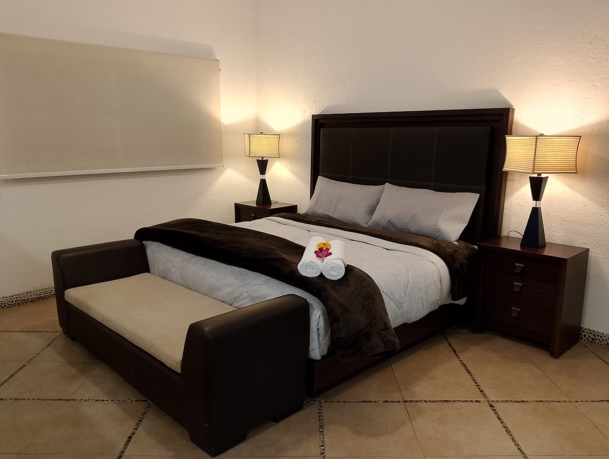 A spacious bed is centered in the room, flanked by two bedside tables with lamps. Neutral tones create a calming ambience, while a soft blanket and rolled towels are arranged thoughtfully on the bed. A large window is fitted with a light-filtering shade.