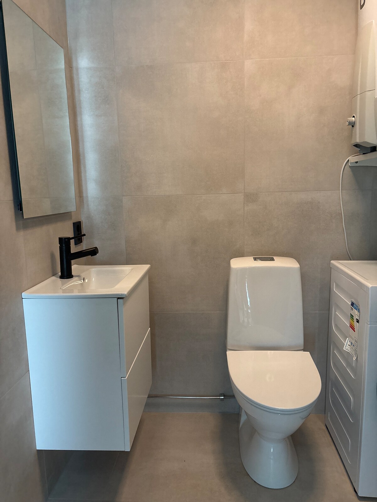 A compact bathroom features modern fixtures, including a sleek toilet and a minimalist sink with a black faucet. A horizontal mirror above the sink reflects the light, while a washing machine is positioned nearby. Neutral tones dominate the tiled walls and floor.