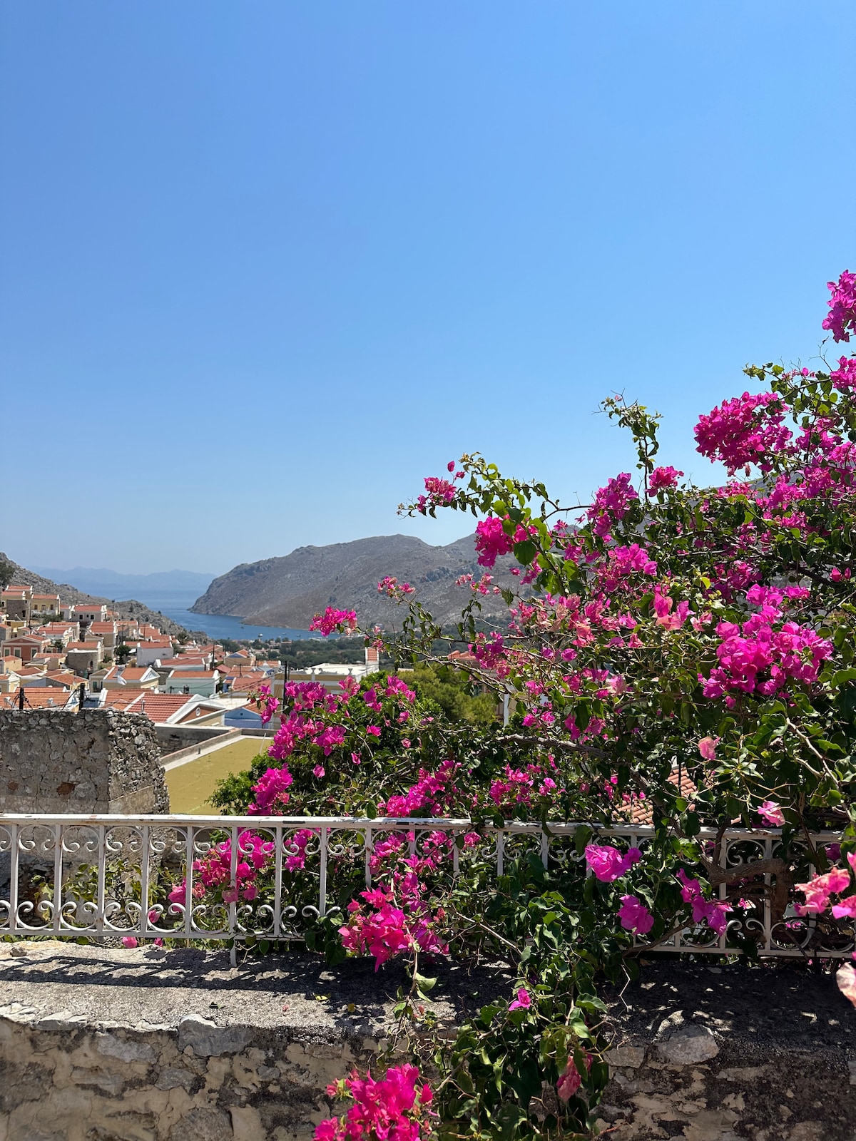 A vibrant display of pink bougainvillea frames the view of the surrounding hills and coastline. The bright flowers contrast with a clear blue sky, while small houses with terracotta roofs are visible in the distance, creating a serene and picturesque scene.