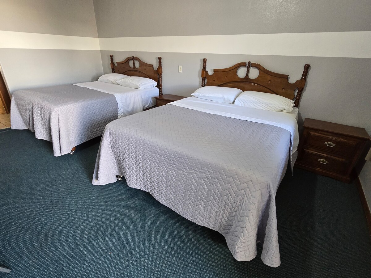 Two spacious beds are positioned in a well-lit room, each adorned with soft gray quilts. Nightstands are placed beside each bed, providing convenient access to essentials. The carpeted floor and neutral wall colors contribute to a calming ambiance.