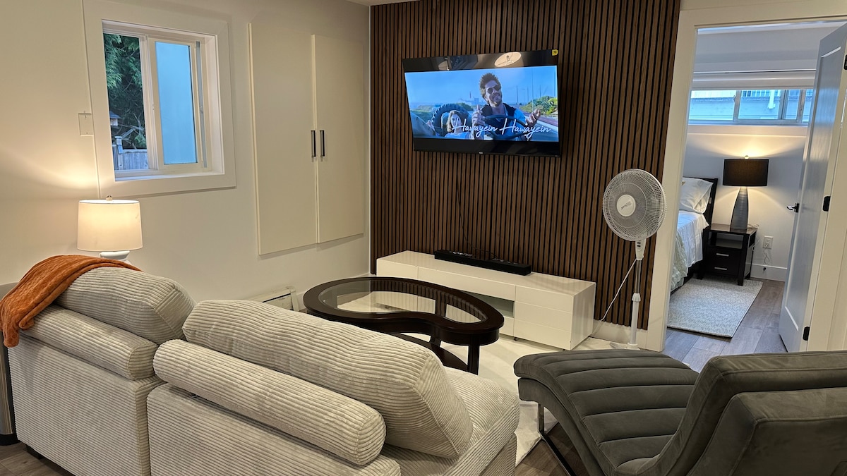 A cozy living area features a light-colored sectional sofa with a textured fabric and contrasting accent throw. A sleek coffee table sits in front, while a wall-mounted television is positioned across from the seating. A fan and lamp provide additional comfort, and a doorway leads to a bedroom.