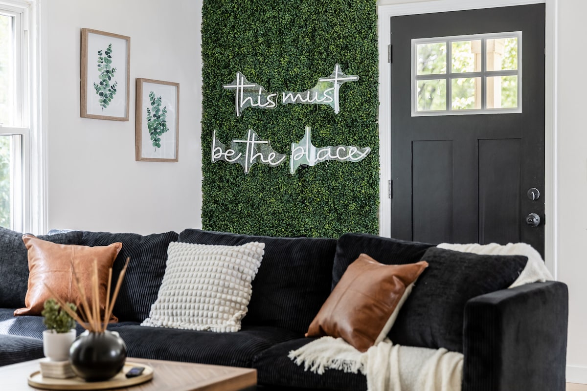 A cozy living area is highlighted by a dark sectional sofa adorned with patterned and solid cushions. A green wall with decorative foliage features a light sign displaying the phrase, 'this must be the place.' Natural light filters in through the front door, enhancing the inviting space.