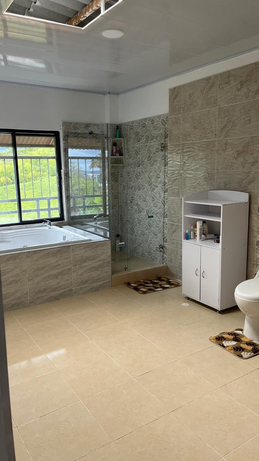 A spacious bathroom features a jetted bathtub next to a large window, providing views of the surrounding landscape. A white cabinet holds toiletries, while a modern toilet is positioned to the side. Light-colored tiles enhance the room's brightness.