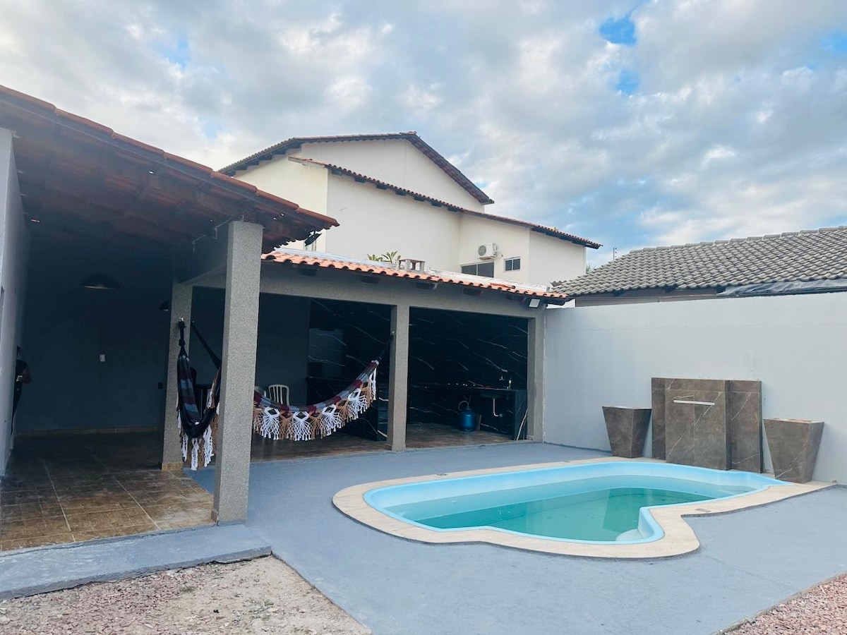 An outdoor area features a uniquely shaped swimming pool with a light blue hue. A covered patio is presented alongside, showcasing a hammock and decorative wall hangings. The background highlights a two-story building under a partly cloudy sky.