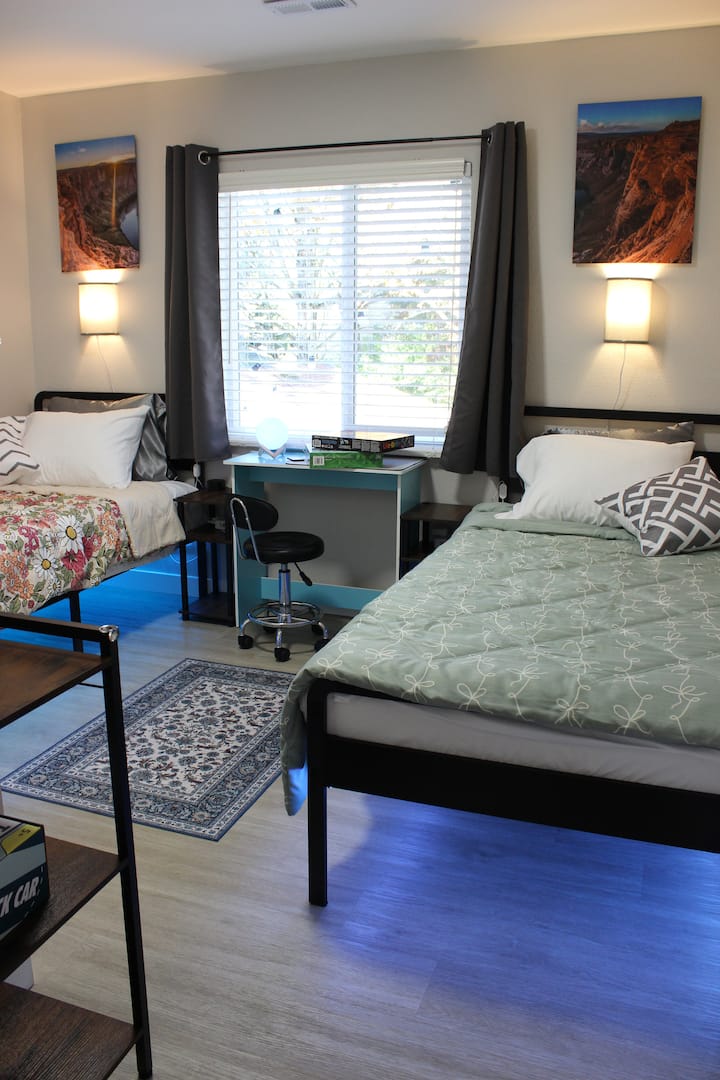 Slaapkamer met twee XL eenpersoonsbedden en een bureau met uitzicht op de grote, omheinde achtertuin, omlijst door volgroeide bomen en gloeiende lichtslingers. Voorzien van LED-kleurveranderende onderbedverlichting, een ruime kast en royale opbergruimte onder het bed.