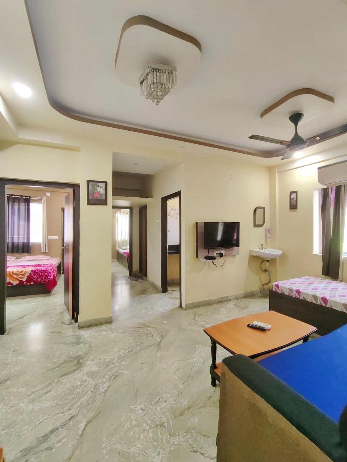 A spacious living area features a television mounted on the wall and a small table with seating. Two bedrooms are visible, showcasing beds with colorful linens. Natural light filters through the curtains, illuminating the marble flooring and highlighting the room's clean design.