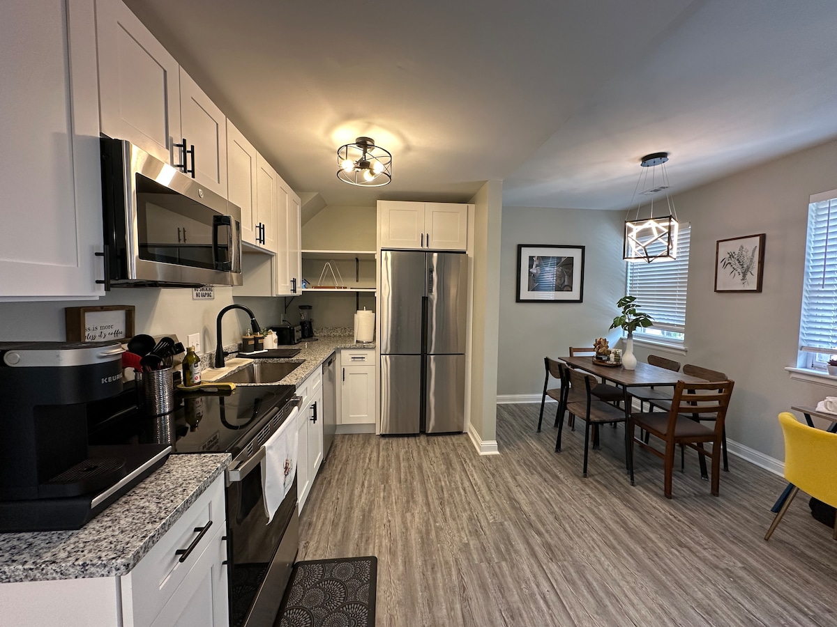 A modern kitchen features sleek white cabinetry and stainless-steel appliances. A granite countertop provides ample preparation space. Adjacent to the kitchen, a dining area is furnished with a wooden table and several chairs, illuminated by a stylish light fixture and natural light from nearby windows.