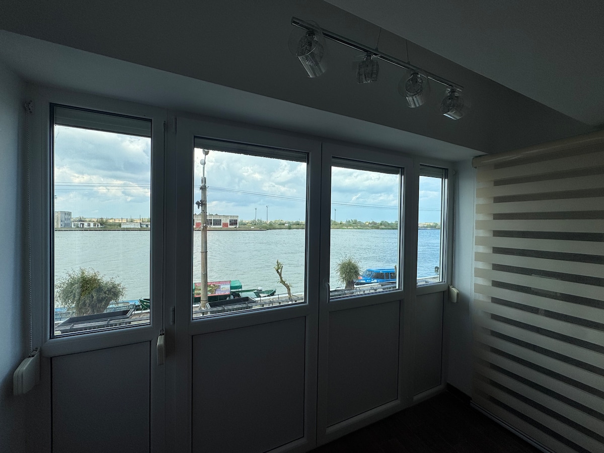 A series of large windows offers a view of the river, framing the tranquil water and distant urban landscape. Natural light illuminates the interior space, complemented by modern ceiling lighting.