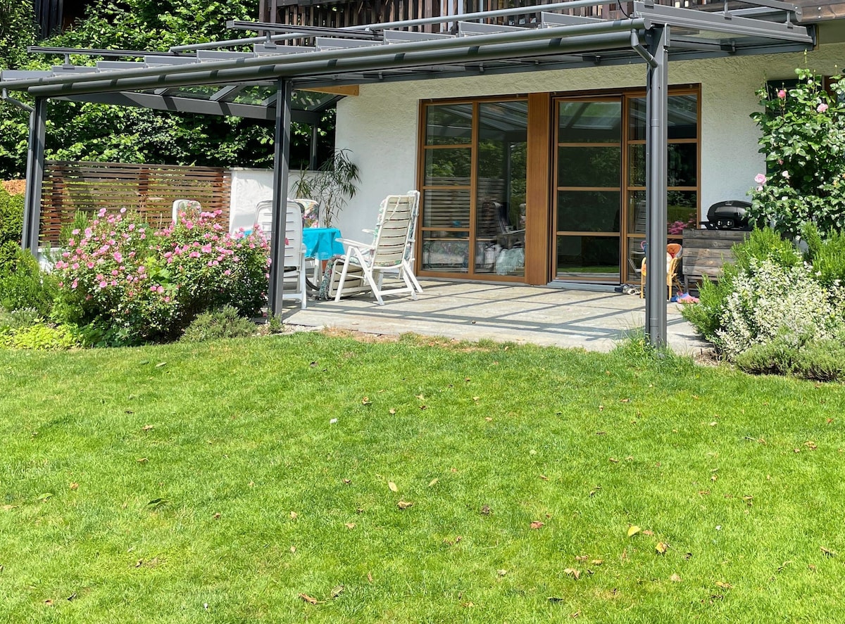 A spacious, covered terrace opens to a well-maintained garden, featuring lush green grass and blooming flowers. The terrace includes a table and several chairs, providing an inviting area for relaxation or dining outdoors. A stylish grill can be seen in the corner.
