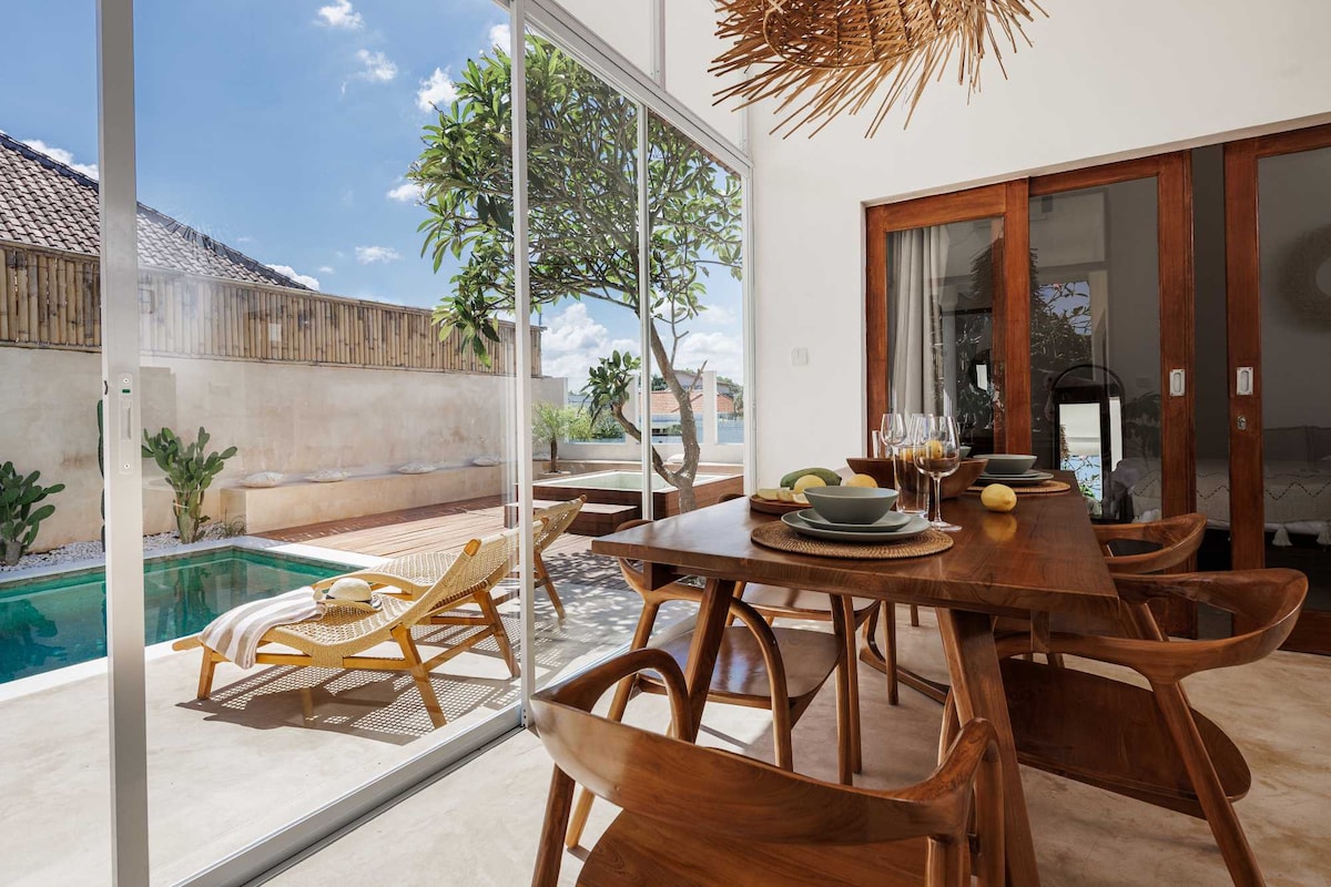 A dining area is presented with a wooden table set for meals, surrounded by modern, curved chairs. Large glass doors open to a serene outdoor space, featuring a swimming pool and lounge chairs under the sun. Natural light brightens the space, complemented by a decorative ceiling fixture.
