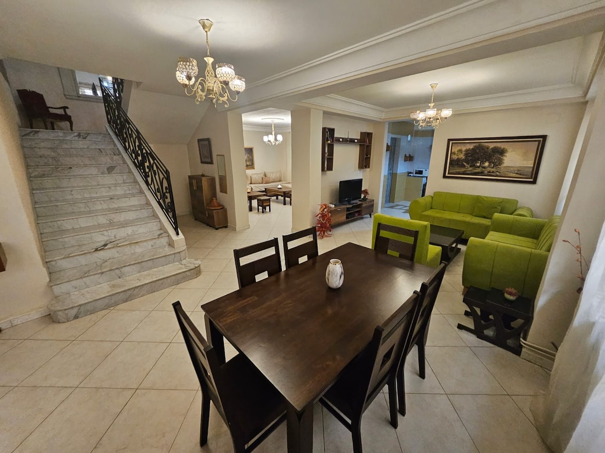 A spacious living area showcases a modern dining table surrounded by black chairs. Bright green sofas are positioned in an inviting arrangement, while a staircase leads to the upper level. Soft lighting from ornate chandeliers enhances the open layout, complemented by light-colored walls and tiled flooring.