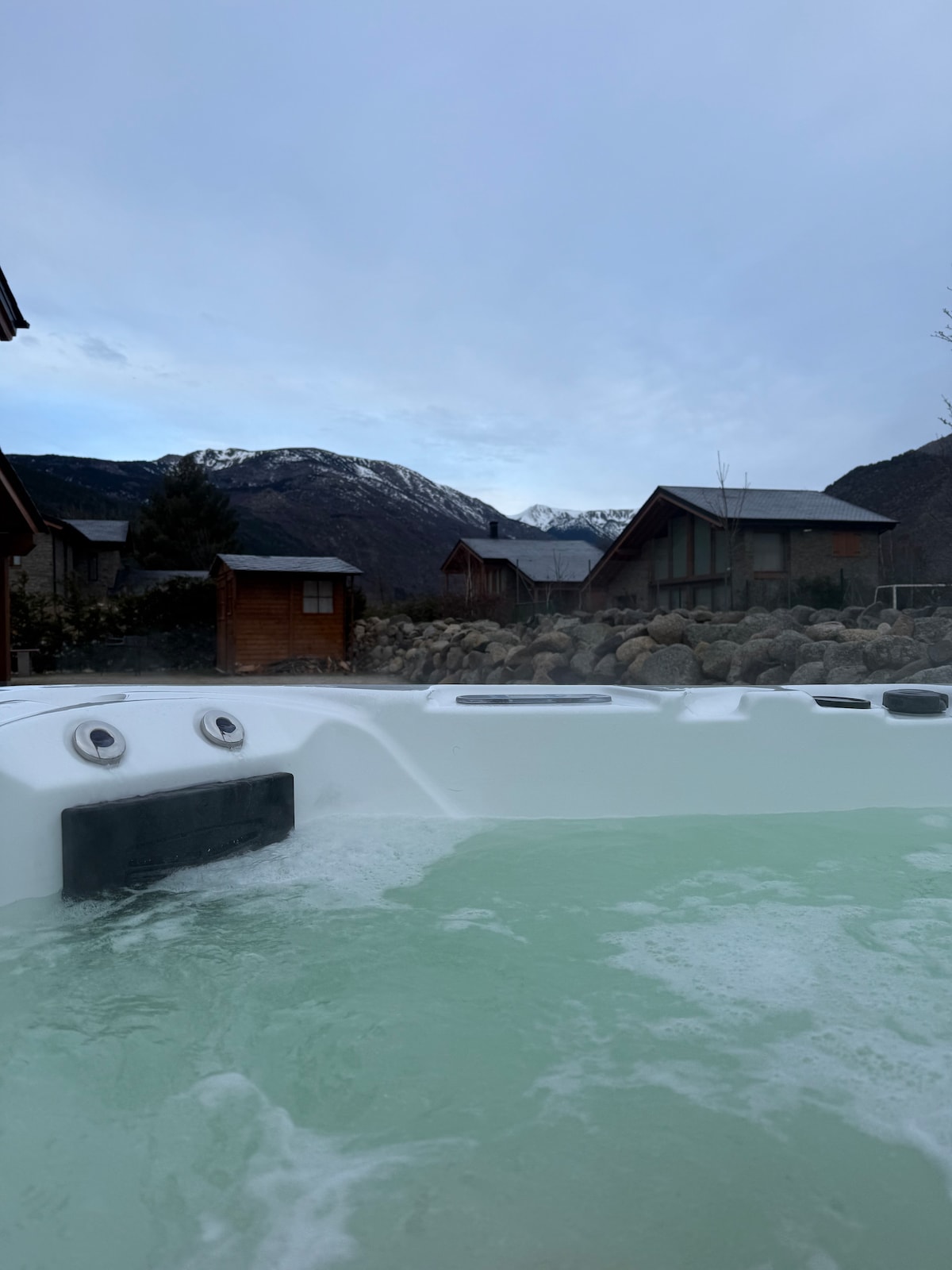 A spacious hot tub is filled with light green water and features jets visible along one side. In the background, wooden cabins are set against a mountainous landscape, showcasing snow-capped peaks under a partially cloudy sky.