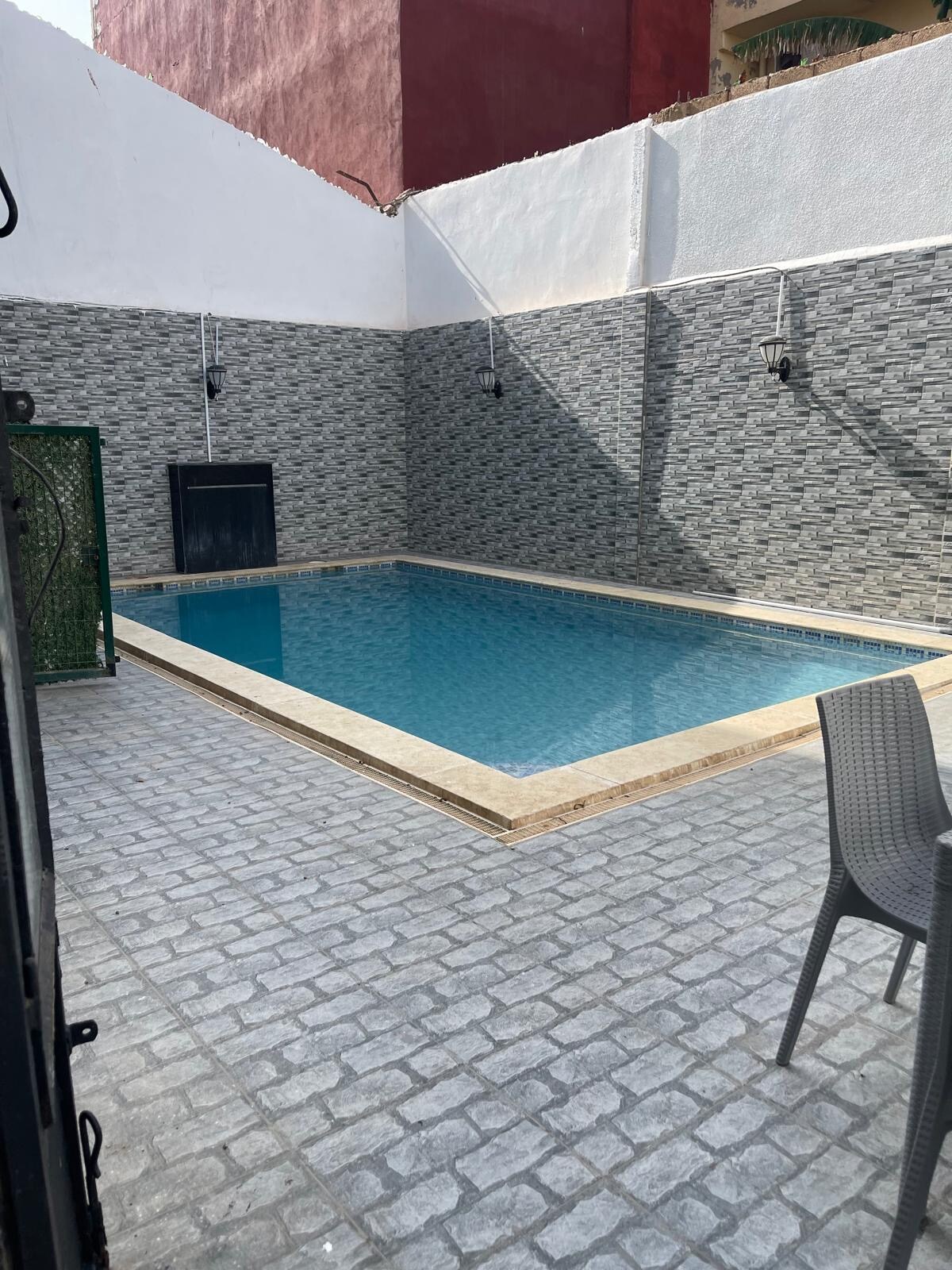 A refreshing swimming pool is surrounded by textured stone walls and laid with patterned pavers. Two wall-mounted lights provide illumination, while a single gray chair sits nearby, offering a convenient seating area. The clear water invites relaxation and leisure.
