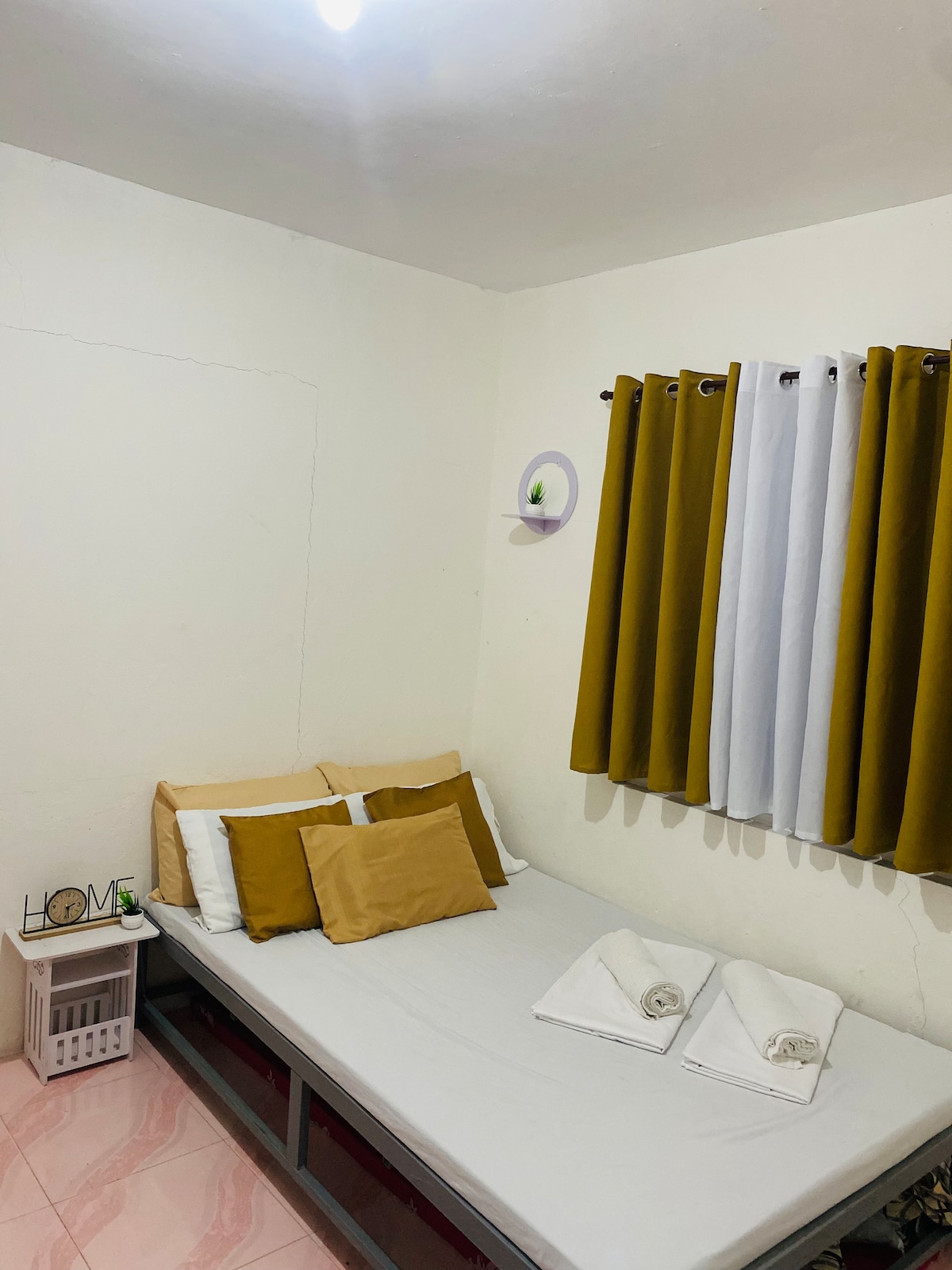 The room features a spacious bed with neutral and mustard-colored pillows. Fresh linens and towels are neatly arranged at the foot of the bed. Yellow and white curtains frame the window, adding a touch of color to the light-colored walls.