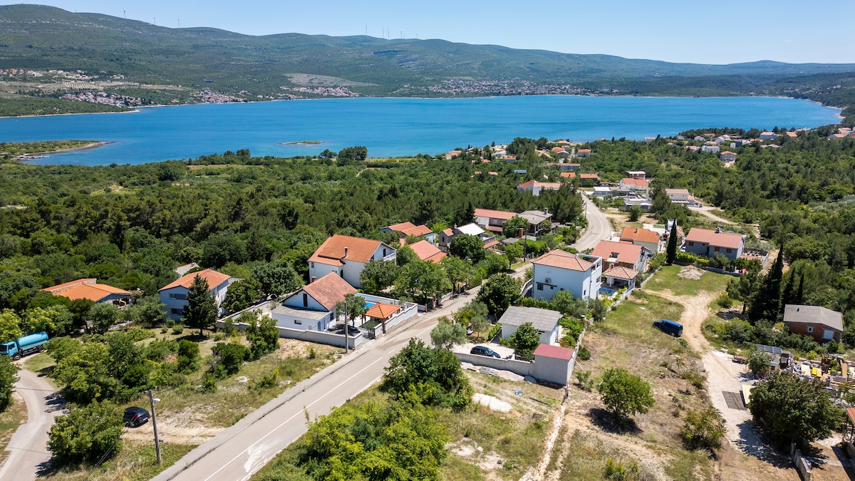 A panoramic view reveals a scenic landscape featuring a calm turquoise bay bordered by lush greenery. Quaint residential homes with terracotta roofs complement the natural surroundings. A winding road leads through the area, enhancing the sense of tranquility within this coastal setting.