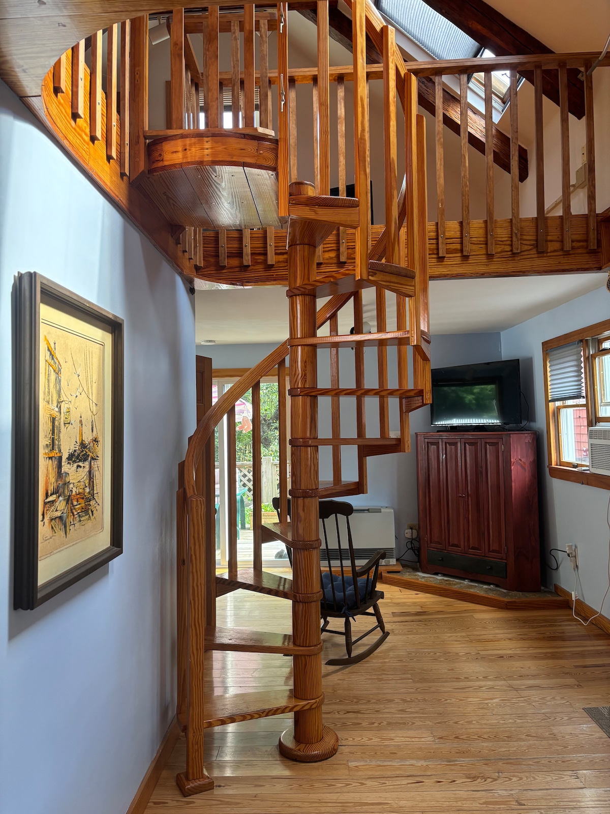 A spiral staircase crafted from rich wood serves as a centerpiece, leading to an upper loft area. Light streams through windows, illuminating the wooden flooring. A framed artwork adorns the wall, while a slim television sits on a cabinet nearby, contributing to the space's functionality.