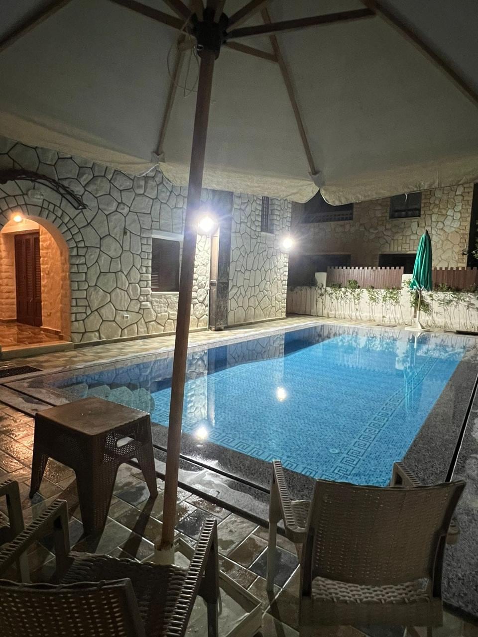 An inviting pool area features a clear blue pool, framed by stone walls. Soft lighting casts a warm glow on the water. Nearby, a shaded seating area includes a table and wicker chairs, creating a comfortable spot for relaxation.