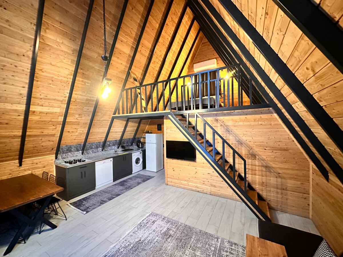 An airy loft-style cabin is highlighted by a wooden A-frame design. A functional kitchen area with modern appliances is situated on the lower level. A cozy staircase leads to an upper sleeping area, with warm lighting creating an inviting environment. A dining table and rug complete the space.