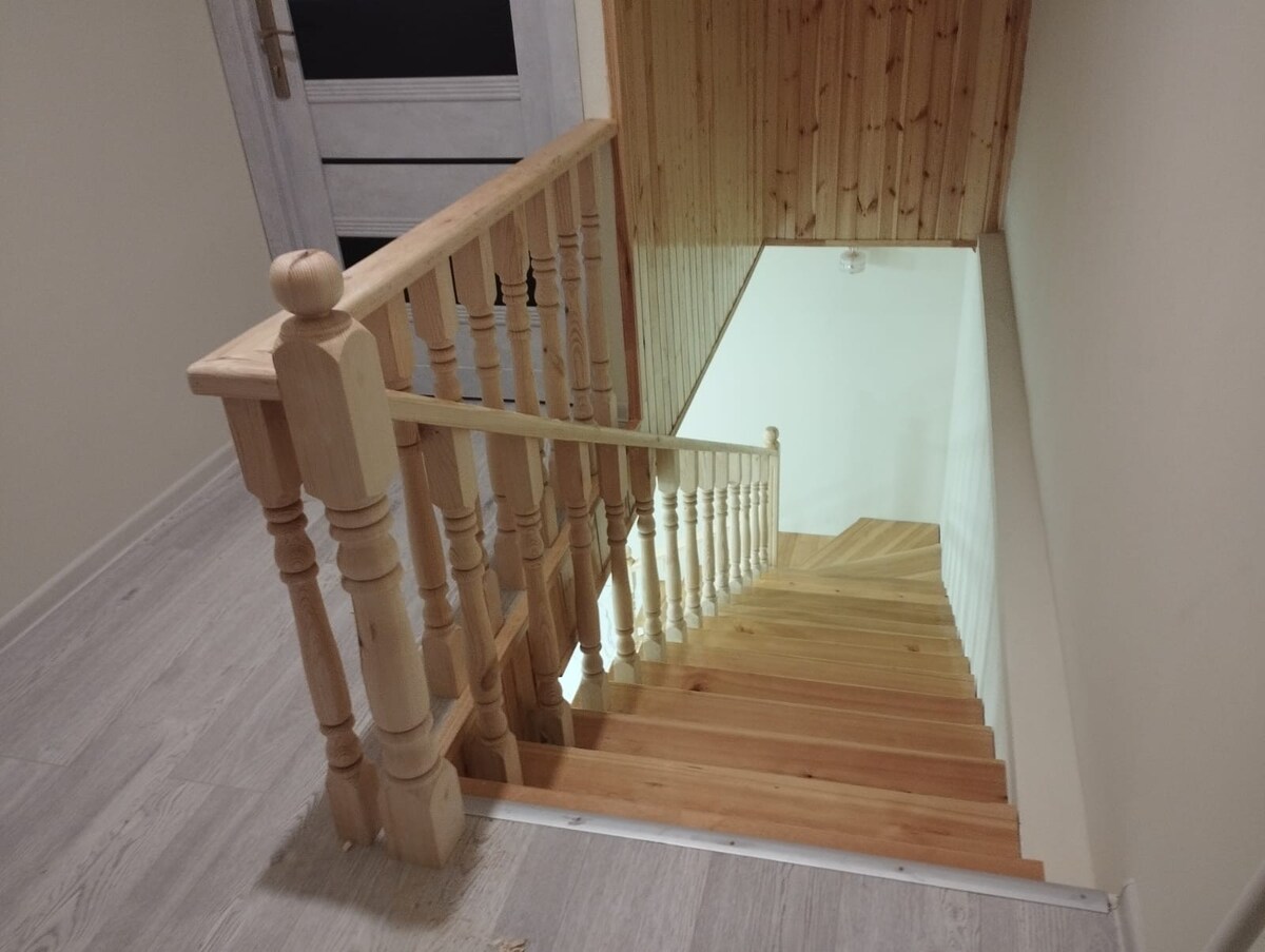 A wooden staircase leads down to a lower level, featuring a light-colored wooden railing and steps. The walls are finished in a neutral tone, enhancing the natural light from above. The flooring displays a modern light wood finish, complementing the overall design.