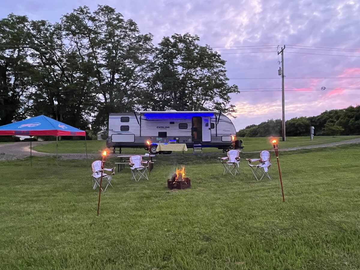 A campsite is presented with a travel trailer, featuring a retractable awning. Four white camping chairs are arranged around a central fire pit, with torches providing ambient light. Lush green grass surrounds the setup, while trees are visible in the background, and a tranquil sky transitions in color.