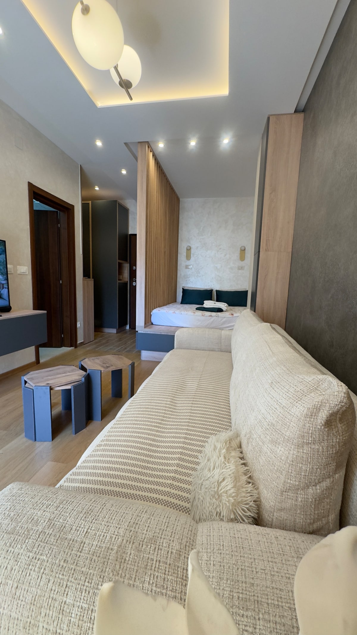 A modern studio space is presented with a cozy sofa featuring textured cushions. The wood-paneled separation divides the living area from the sleeping space, where a spacious bed is subtly visible. Soft lighting complements the neutral color palette, creating a warm ambiance.