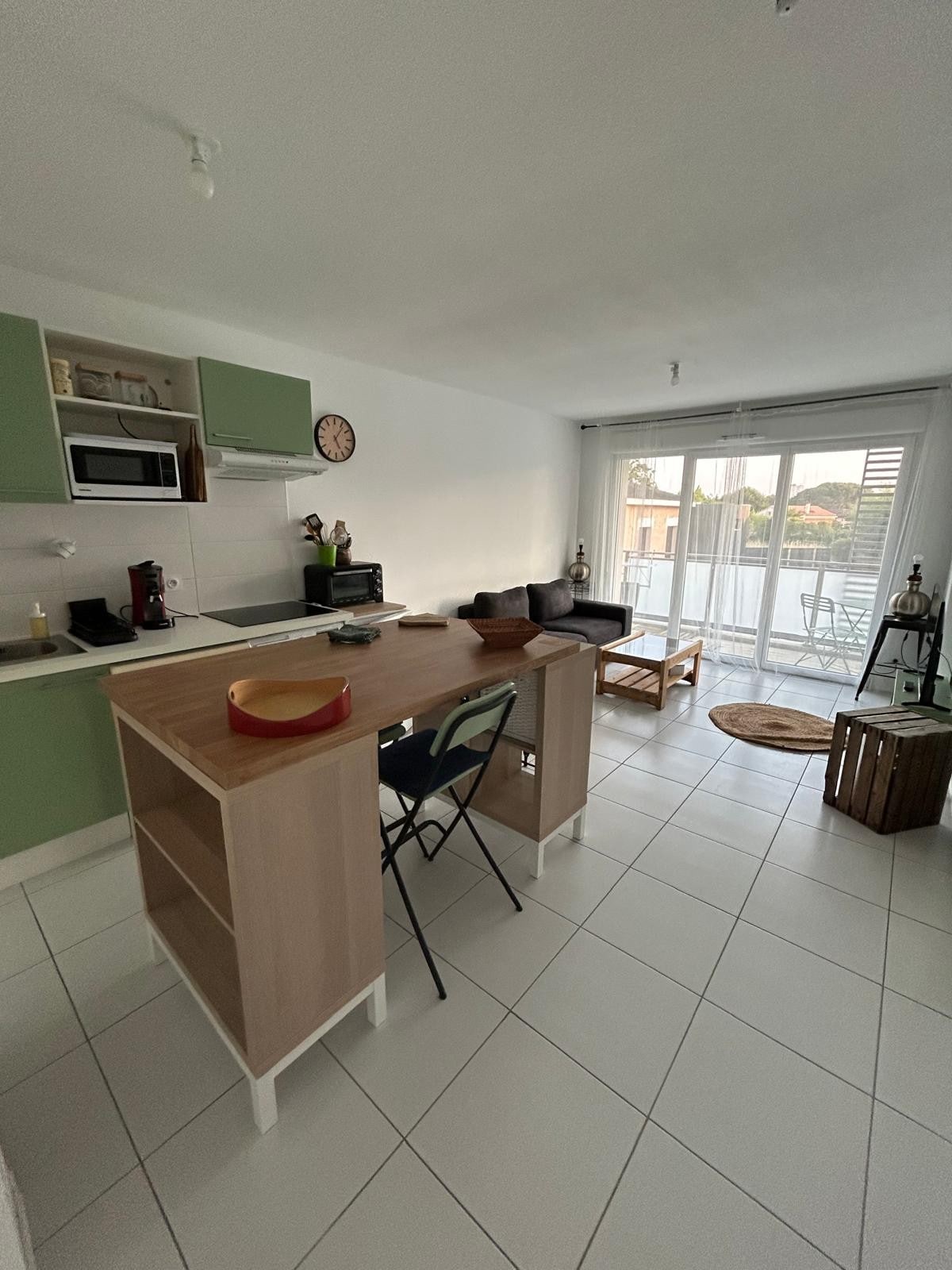 The open-concept living space features light-colored tiled flooring. A contemporary kitchen with green cabinetry is seen, alongside a wooden dining table and two chairs. A comfortable seating area with a sofa and coffee table is positioned beside large glass doors leading to a balcony.