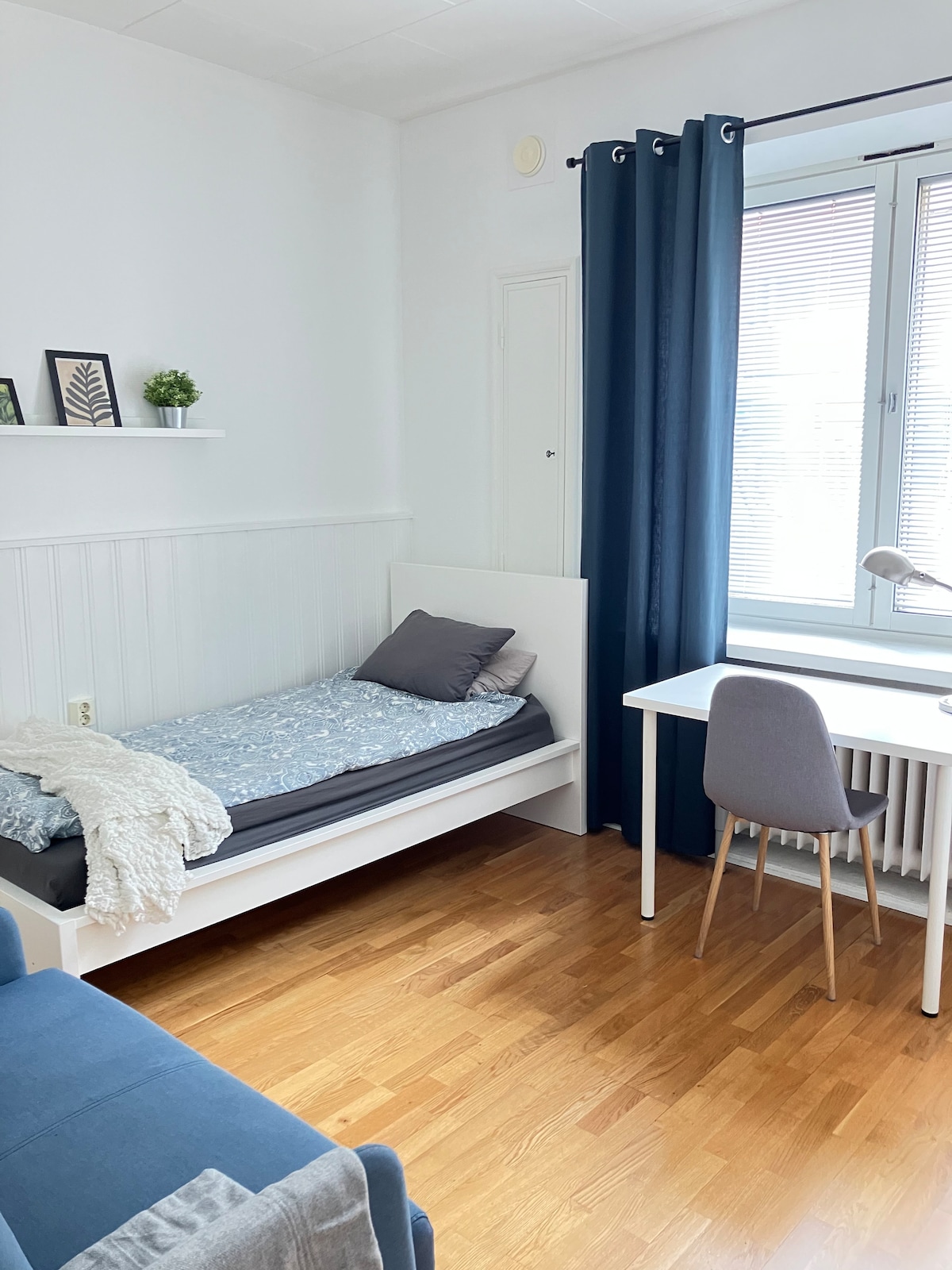 A sleeping area is furnished with a single bed, featuring a light blue and gray patterned duvet. A desk with a chair and a small blue sofa are arranged along the wall. Natural light filters through a window dressed with dark curtains, illuminating the wooden floor.