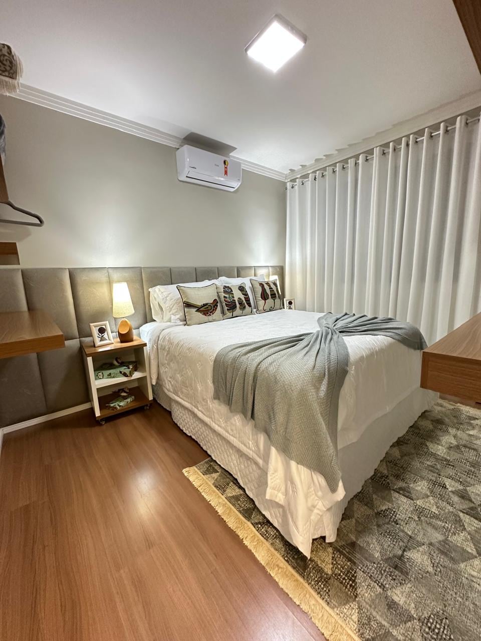 A cozy bedroom features a spacious bed adorned with decorative pillows and a soft blanket. Natural light filters through sheer curtains, creating a bright atmosphere. A small bedside table holds a lamp and personal items, while a stylish rug adds warmth to the wooden floor.