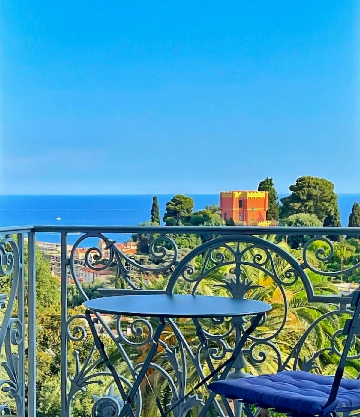 A charming balcony features a circular table surrounded by elegant wrought iron railings. The view overlooks the Mediterranean Sea, with lush greenery and a historic building in the background, highlighting the scenic coastal landscape.