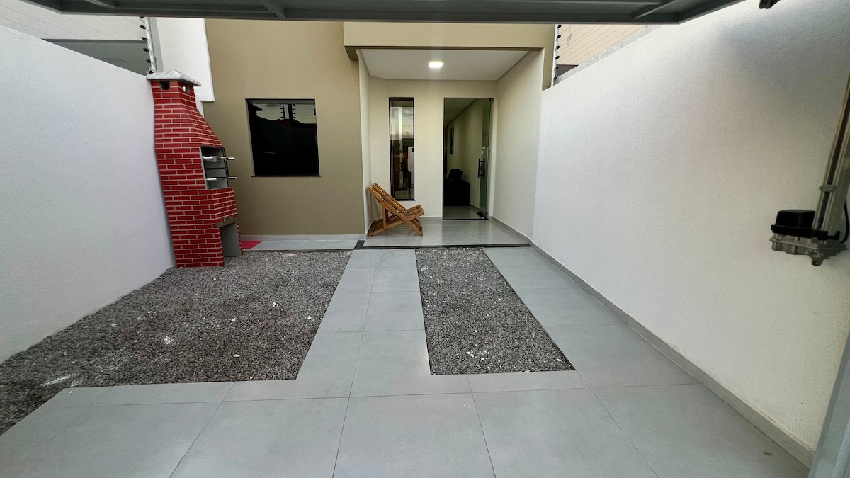 An outdoor area features a modern barbecue grill with a brick facade. The flooring is a combination of large gray tiles and gravel patches, creating a clean and open space. The entrance to a well-lit interior area is visible in the background.