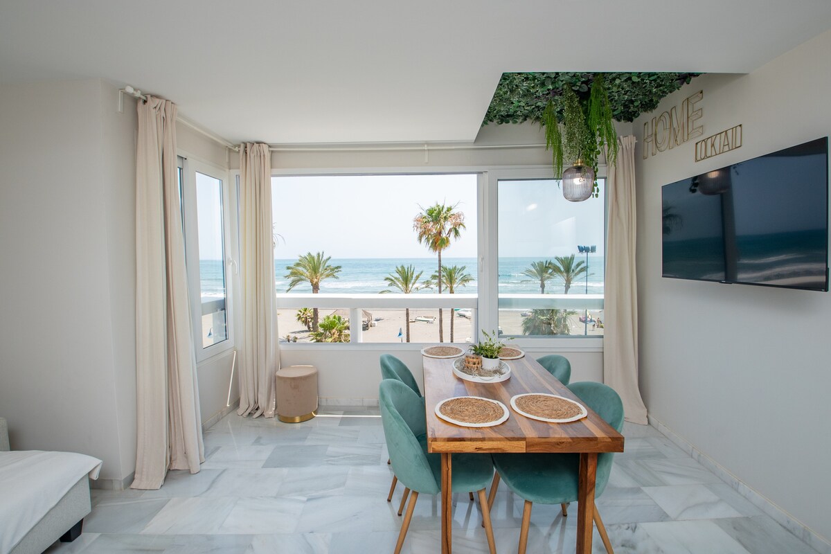 A bright dining area features a wooden table surrounded by soft green chairs. Large windows offer expansive views of the beach and palm trees, allowing natural light to fill the space. White walls and a flat-screen TV enhance the modern aesthetic.