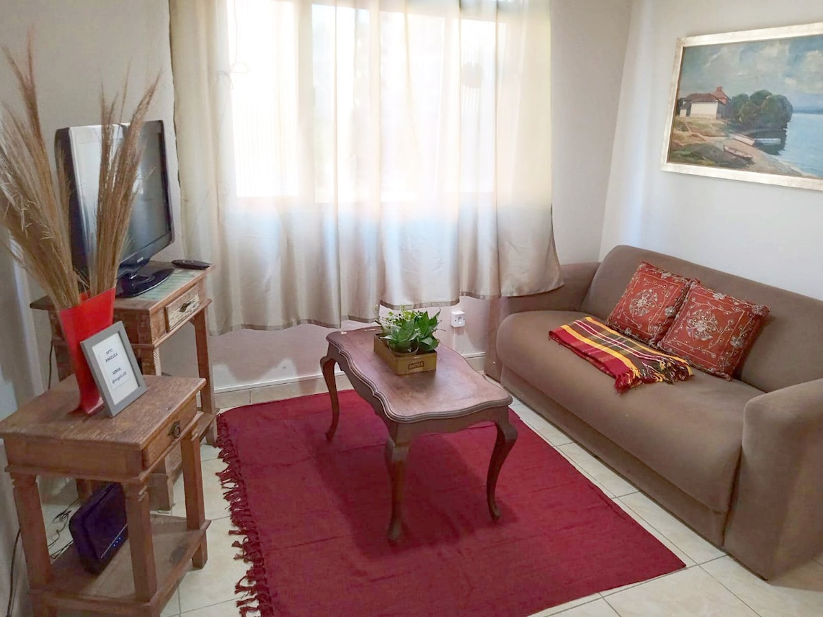 A living area is highlighted by a comfortable sofa adorned with decorative cushions. A coffee table is set in front, complemented by a potted plant. Natural light filters in through sheer curtains, and a television is positioned nearby, enhancing the cozy atmosphere.