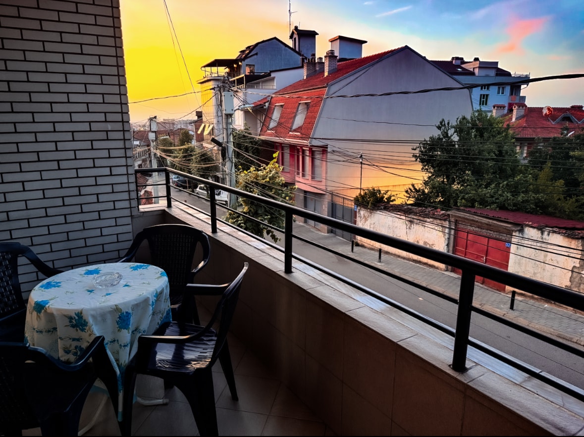 A private balcony is showcased, featuring a round table with a floral tablecloth and four black chairs. The view reveals a sunset sky with warm colors, while the surrounding buildings and greenery are visible in the background.