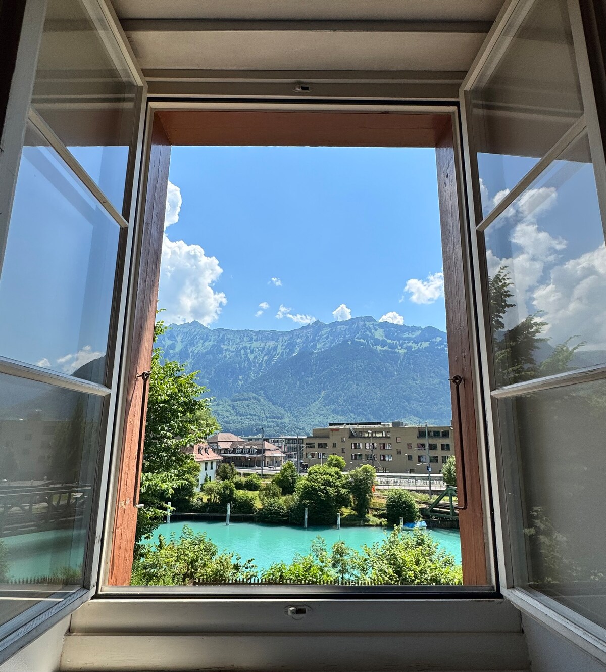 A scenic view is framed by open windows, showcasing the vibrant turquoise waters of the Aare River. Lush trees line the riverbank, while the impressive Jungfrau mountain range rises majestically in the background under a clear blue sky. Fluffy white clouds complete this captivating panorama.