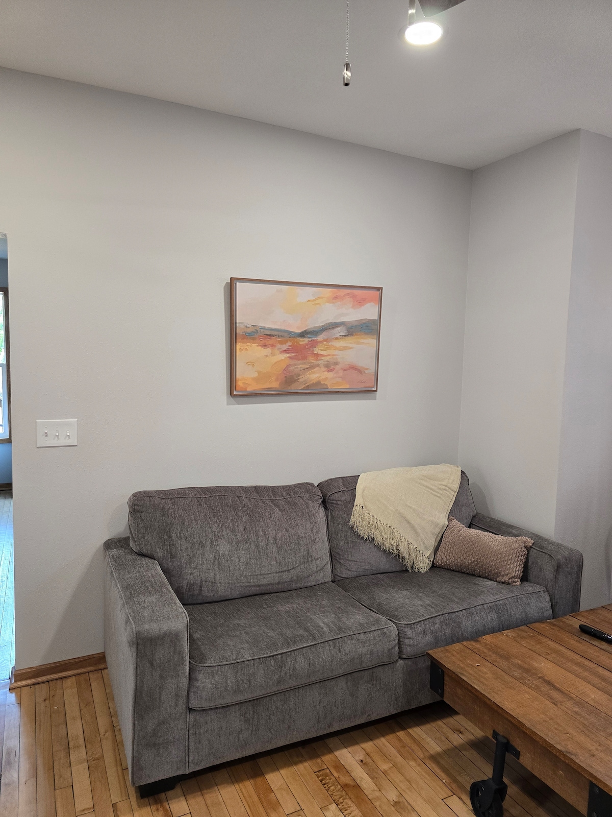 A comfortable gray sofa is positioned against a light-colored wall. A muted artwork is displayed above the sofa, adding a touch of color. A textured throw blanket drapes casually over one arm, while a decorative pillow complements the seating area.
