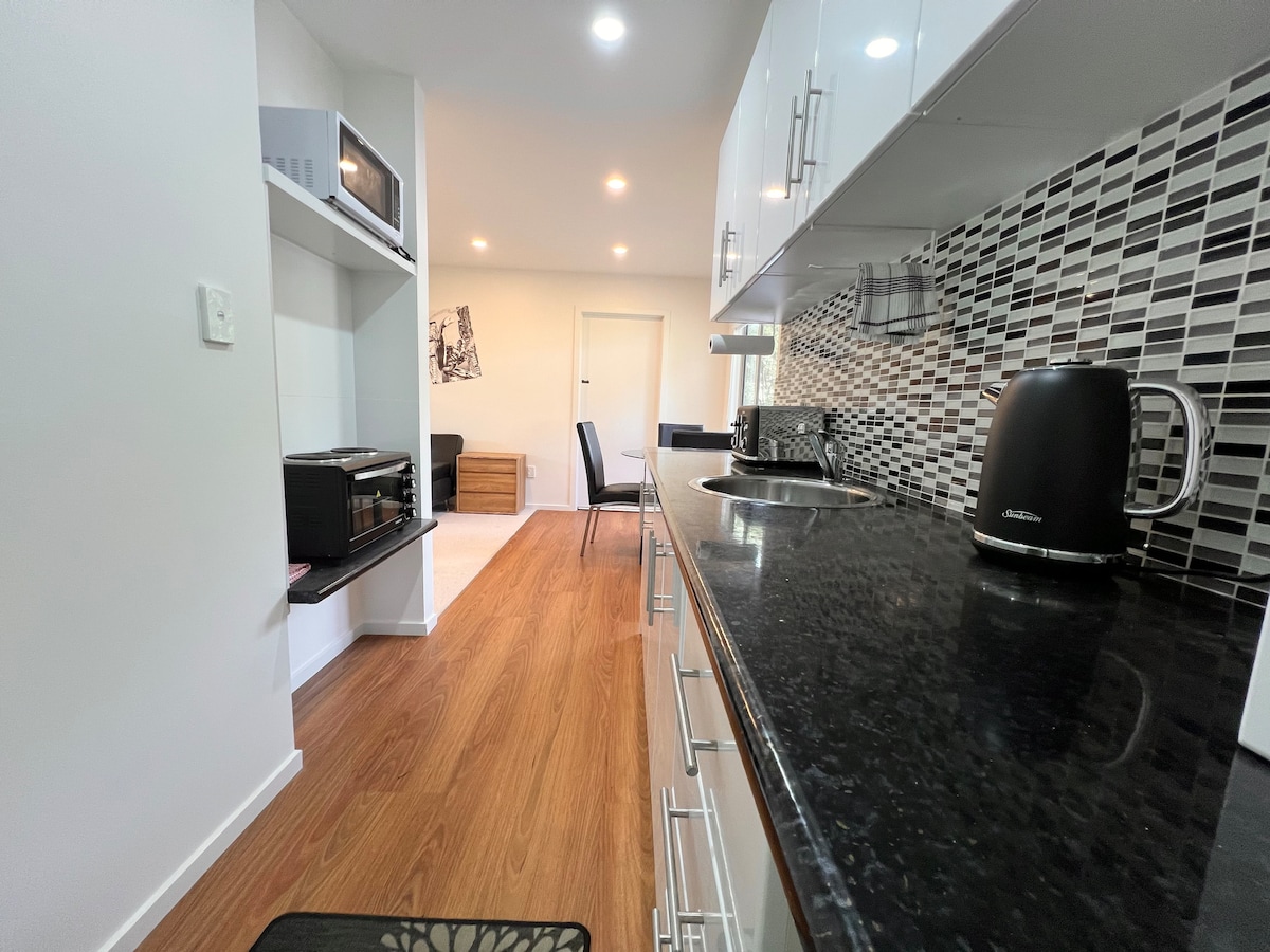 A modern kitchenette is presented with sleek black countertops and a geometric backsplash. Essential appliances, including a microwave and kettle, are visible. The open layout extends to a dining area featuring a simple table and chair set, with natural light enhancing the space.