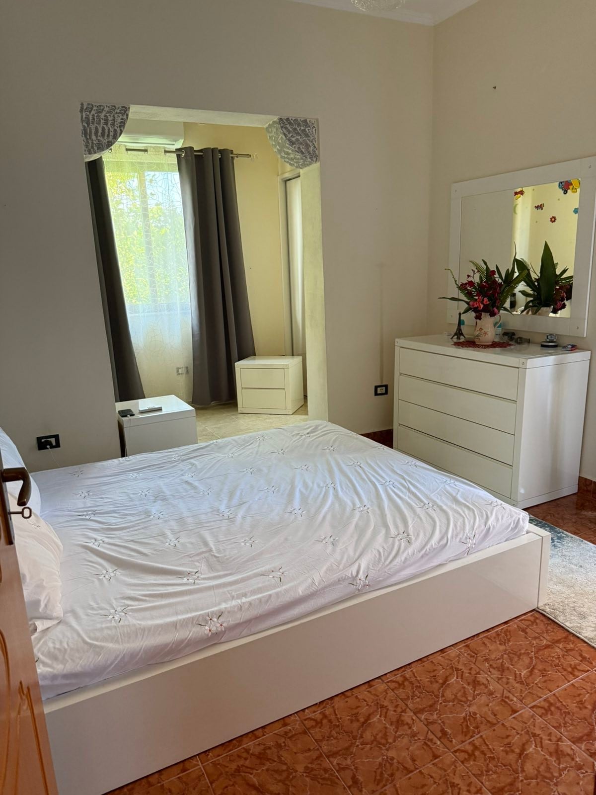 A sleeping area features a spacious bed with a simple white coverlet. A decorative mirror reflects the room's light, enhancing the sense of space. A dresser and two nightstands provide storage, while graceful curtains frame the window, allowing soft light to enter.