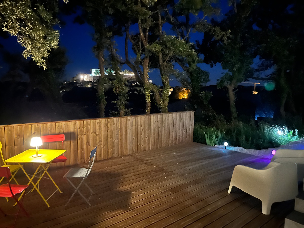 An outdoor terrace is illuminated at night, featuring a small yellow table with four chairs and two lounge chairs. Surrounding trees provide privacy, while a distant view of the Grignan castle is visible against the night sky.