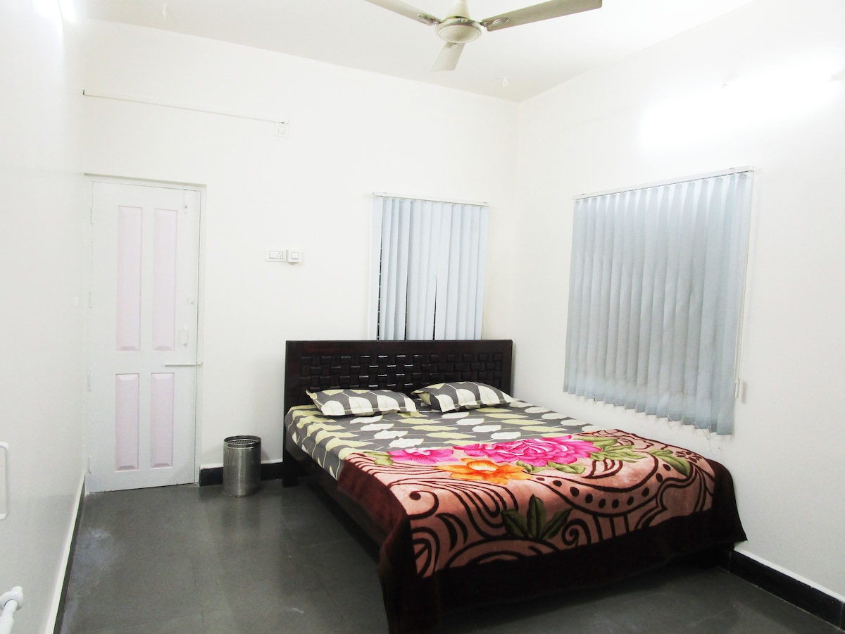 A spacious bedroom is presented with a large bed covered in a patterned blanket featuring floral designs. Two light-colored windows with sheer curtains allow natural light to fill the room. A simple wooden door is visible, along with a small waste bin on the floor.