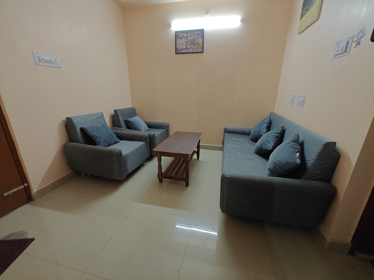 A simple seating area is arranged with two armchairs and a comfortable couch, all in a muted gray fabric. A wooden coffee table is centered in front of the seating. Soft lighting from the ceiling fixture creates a warm ambiance.