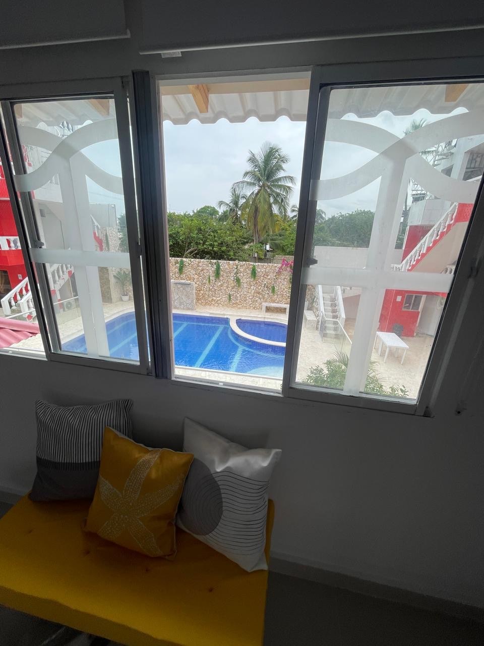 The view showcases a swimming pool framed by palm trees and manicured landscaping. Two large windows provide ample natural light and an unobstructed view of the inviting pool area. Soft pillows in varied colors are arrayed on a bright yellow seat, enhancing the sense of comfort.