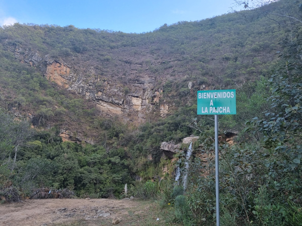 A welcome sign, reading 'Bienvenidos a La Pajucha,' is positioned in front of a natural landscape. The backdrop features a hillside covered in green foliage and rocky terrain under a partly cloudy sky. A small waterfall can be glimpsed among the lush vegetation.