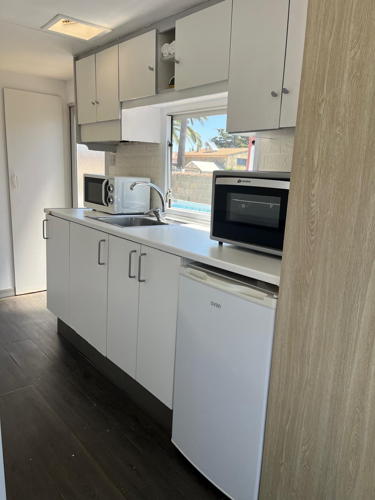 A modern kitchen area features sleek white cabinetry and a countertop with a sink. Appliances include a microwave and a compact refrigerator. Natural light enters through a window, illuminating the dark wood flooring throughout the space.