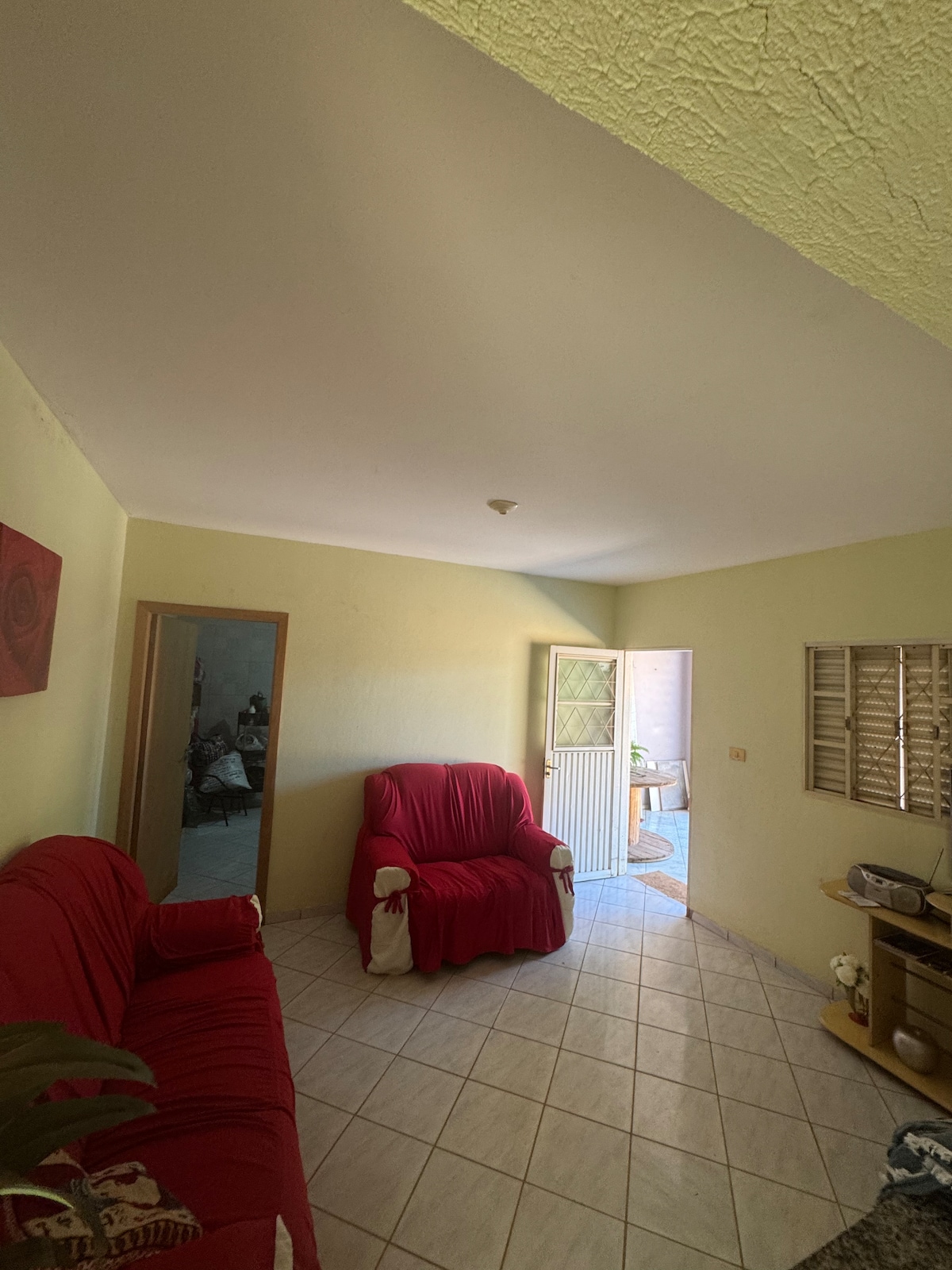 A bright living area features two red armchairs positioned on a tiled floor. Natural light enters through a doorway that leads to an outdoor space. A mirror reflects part of the room, enhancing the sense of openness.