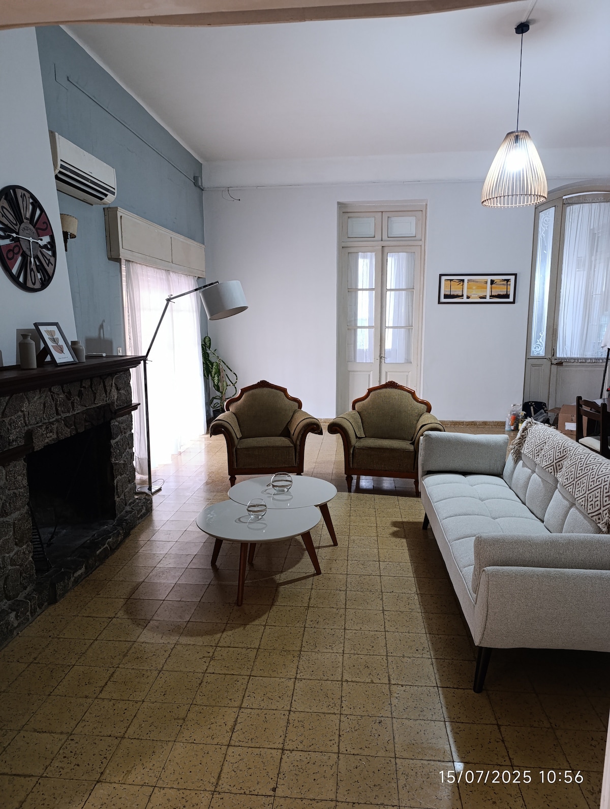 A spacious living area features a comfortable sofa and two vintage armchairs arranged around a coffee table. A stone fireplace is positioned against one wall, and large windows allow natural light to fill the room, enhancing the inviting atmosphere.