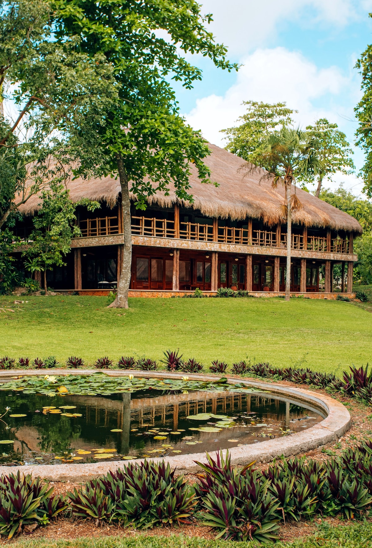 A large, thatched-roof building is surrounded by lush greenery and tall trees. A circular pond reflects the structure and the vibrant landscaping, featuring ornamental plants and grasses along the edge.