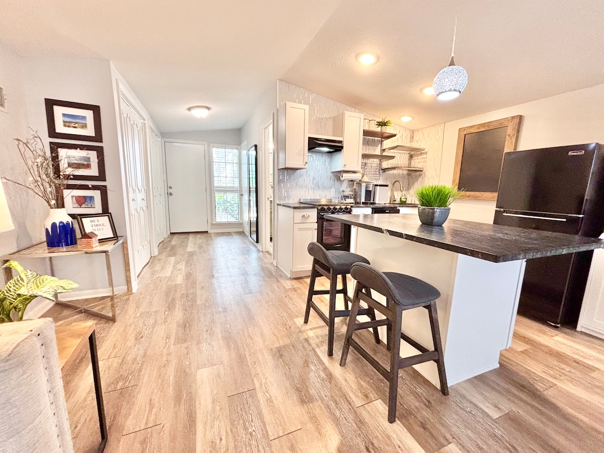 The open concept kitchen features a bar with two stools and modern appliances, including a black refrigerator and stovetop. Natural light filters in through a nearby glass door, highlighting the wood-look flooring and neutral color palette throughout the space.