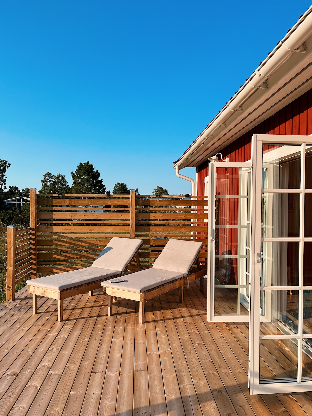 A spacious wooden deck is highlighted, featuring two sun loungers positioned for relaxation. Clear glass doors provide access to the interior, complementing the warm wooden tones of the decking and surrounding fence. Bright blue sky is visible above, enhancing the outdoor ambiance.