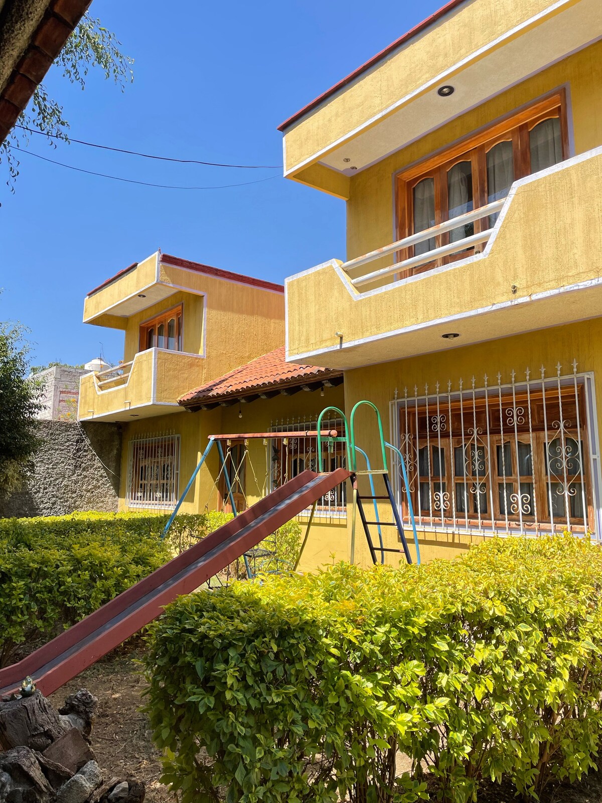A vibrant yellow building is featured, showcasing two levels with large windows adorned with decorative grills. A green slide is positioned in front, set against a backdrop of well-maintained hedges, with clear blue skies overhead enhancing the outdoor play area.
